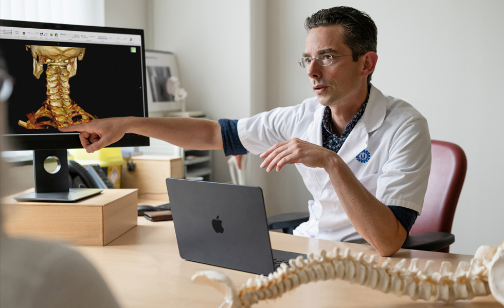 A medical professional points to a 3D skeletal rendering of the back of a neck on a Studio Display, while a MacBook sits on the desk