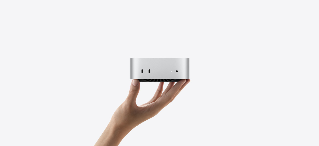 لقطة أمامية لجهاز Mac mini مسنود على رؤوس أصابع يد ممدودة للأعلى، الجهة الأمامية تعرض اثنين من منافذ ثندربولت، ضوء مؤشر التشغيل ومقبس لسماعات الرأس، قاعدة سوداء مدببة في الأسفل، سطح مسطح، جوانب مدورة، حواف مستقيمة، لون فضي