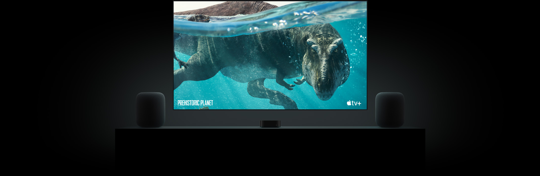 Large flat screen TV featuring a vivid image of a dinosaur from Prehistoric Planet. The TV is hung above an Apple TV and framed by two HomePod speakers placed on a livingroom console