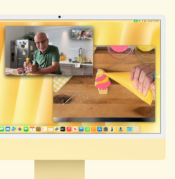 iMac jaune. Démonstration de la fonctionnalité Vue du bureau. Écran montrant une pâtissière lors d’un appel vidéo avec une autre personne. Un écran montre la pâtissière et le visage d’un homme. Un autre écran montre les mains de la pâtissière mettant du glaçage sur un biscuit.