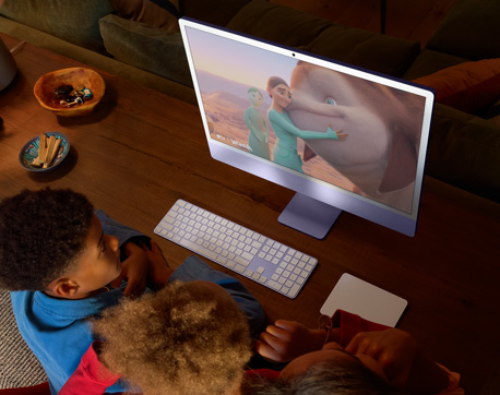 Três jovens sentados à mesa com um iMac, Magic Keyboard e Magic Trackpad, assistindo a um filme