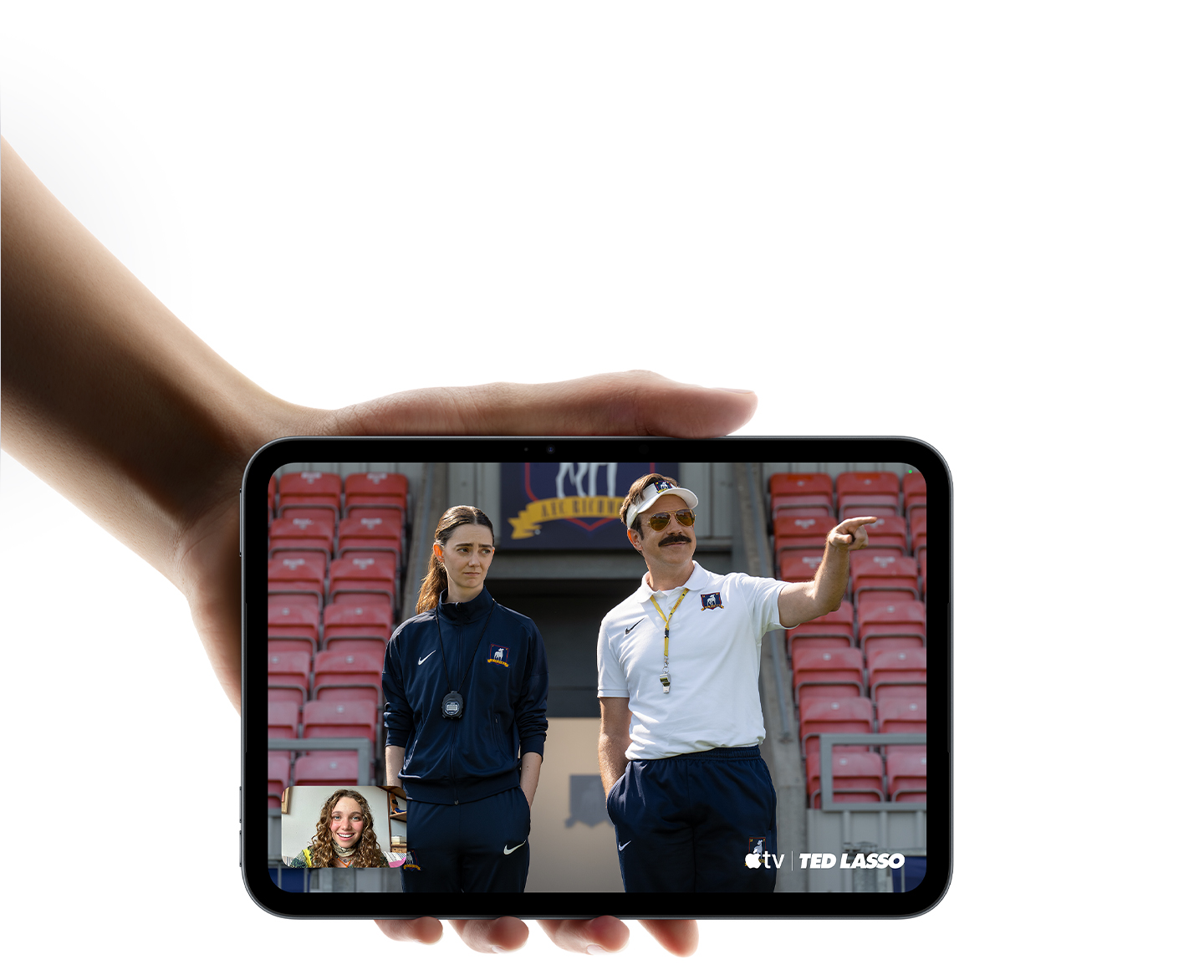 iPad mini in Midnight colour, being held by a hand, displaying Apple TV Show, Ted Lasso being watched with SharePlay on a FaceTime call.