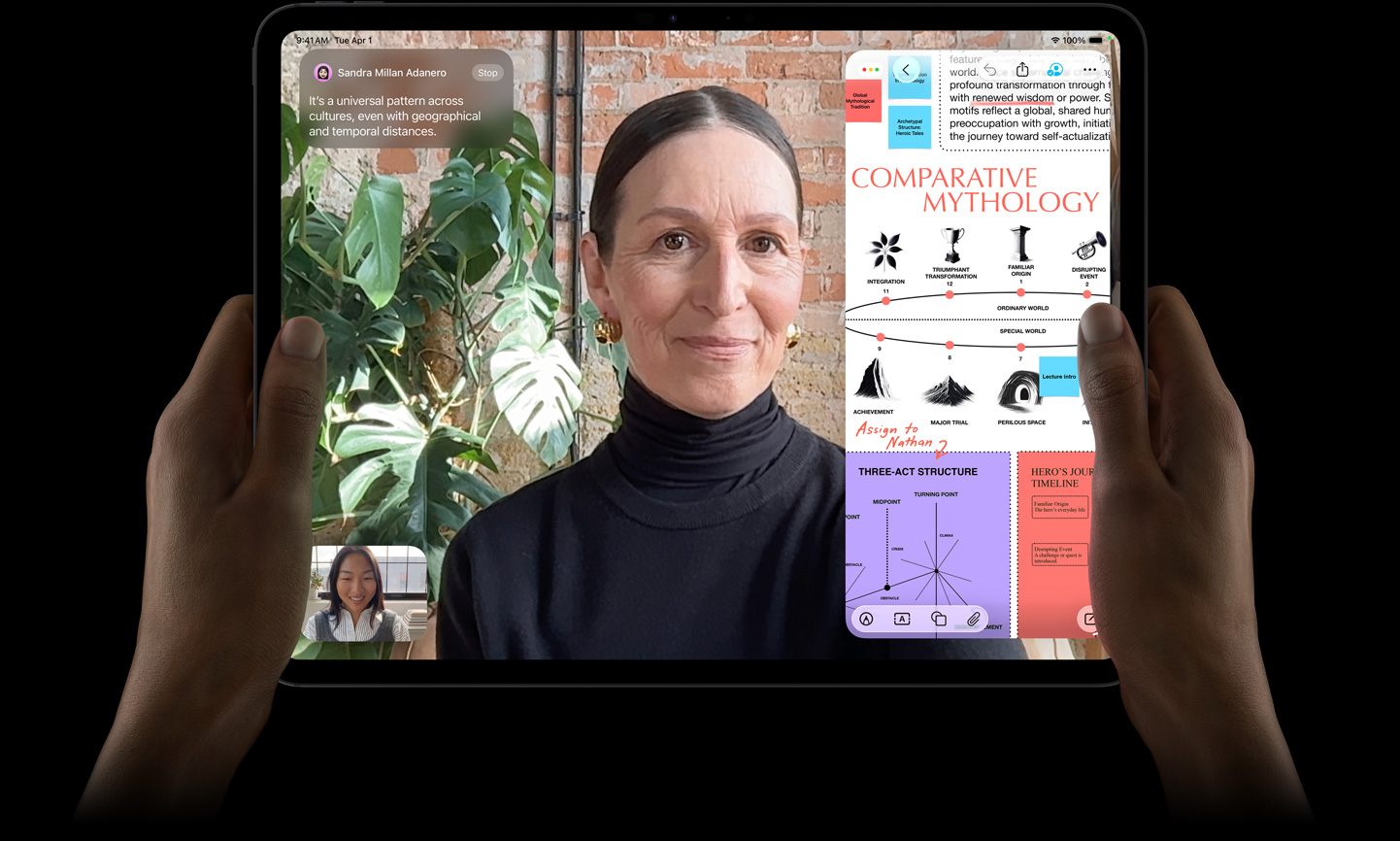 太空黑 iPad 機身正面，左右手以橫向握持，展示圓角設計，黑色顯示器邊框，顯示器顯示正在實時翻譯一段 FaceTime 對話，以及一個教育 app 展示「Comparative Mythology」簡報，其中有文字、圖表和手寫筆記