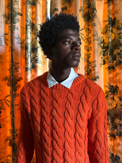 A photo of a man in a bright red sweater standing in front of patterned curtains. The photo was taken in low-light on the Main camera.