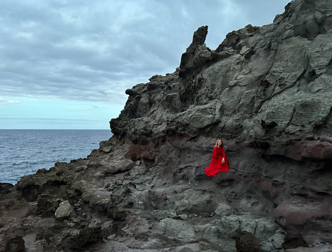 Pääkameralla hämärässä valaistuksessa otettu kuva puna­mekkoisesta naisesta, joka poseeraa kallio taustanaan.