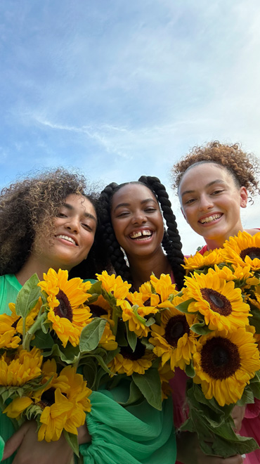 Selfie tajam dan cemerlang tiga orang yang memegang bunga berwarna kuning, diambil dengan kamera TrueDepth