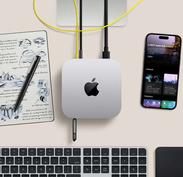 Mac mini, top, silver aluminium, rounded corners, glossy black Apple logo centred, connected by cord, USB stick inserted in USB-C port in front, Studio Display stand partially visible, iPhone, Magic Trackpad, Magic Keyboard with Touch ID, stylus and e-ink sketch pad