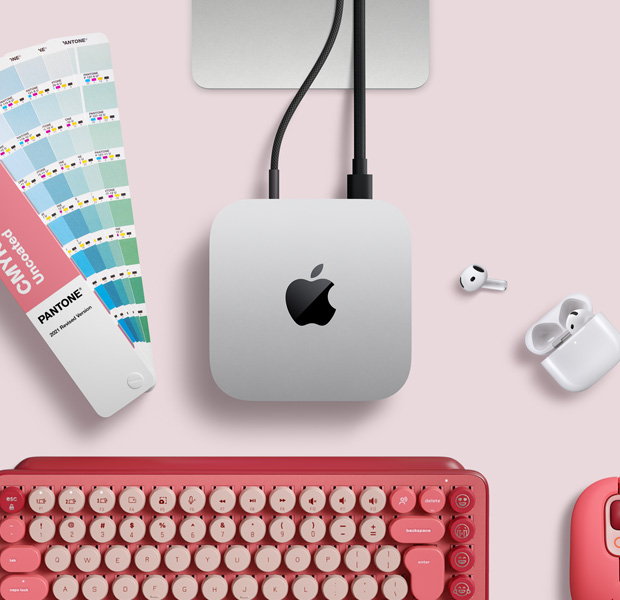 Mac mini, top, silver aluminium, rounded corners, glossy black Apple logo centred, connected by cord, Studio Display stand partially visible, AirPods 4, Pantone colour swatches, and red Logitech keyboard and mouse