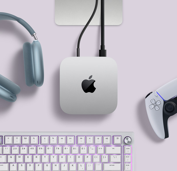 Mac mini, top, silver aluminium, rounded corners, glossy black Apple logo centred, connected by cord, Studio Display stand partially visible, AirPods Max 2, blue colour, PlayStation DualSense controller, white colour and white keyboard