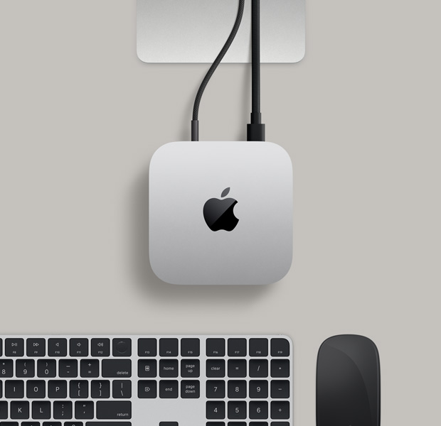 Mac mini, top, silver aluminium, rounded corners, glossy black Apple logo centred, Studio Display stand partially visible, Magic Keyboard with Touch ID, Magic Trackpad