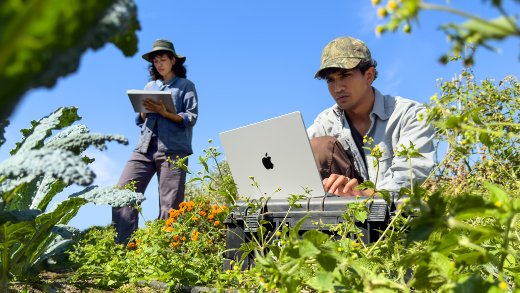 Duas pessoas trabalhando ao ar livre em um jardim. Uma delas usa um MacBook Pro prateado desconectado da tomada, e a outra segura um bloco de notas