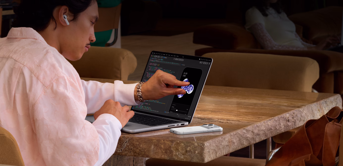 A person sits at a table in a public space, they use a pinch gesture to adjust an element in Xcode using the Touch feature of a silver MacBook Pro with PROLED XDR Display, an iPhone is on the table beside the laptop and the person is using AirPods Pro