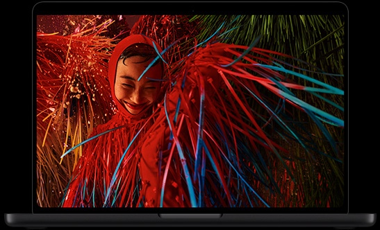 MacBook Pro with PROLED XDR Display, space black colour, open, display shows a photo of a smiling person in a surreal red outfit with feathery details